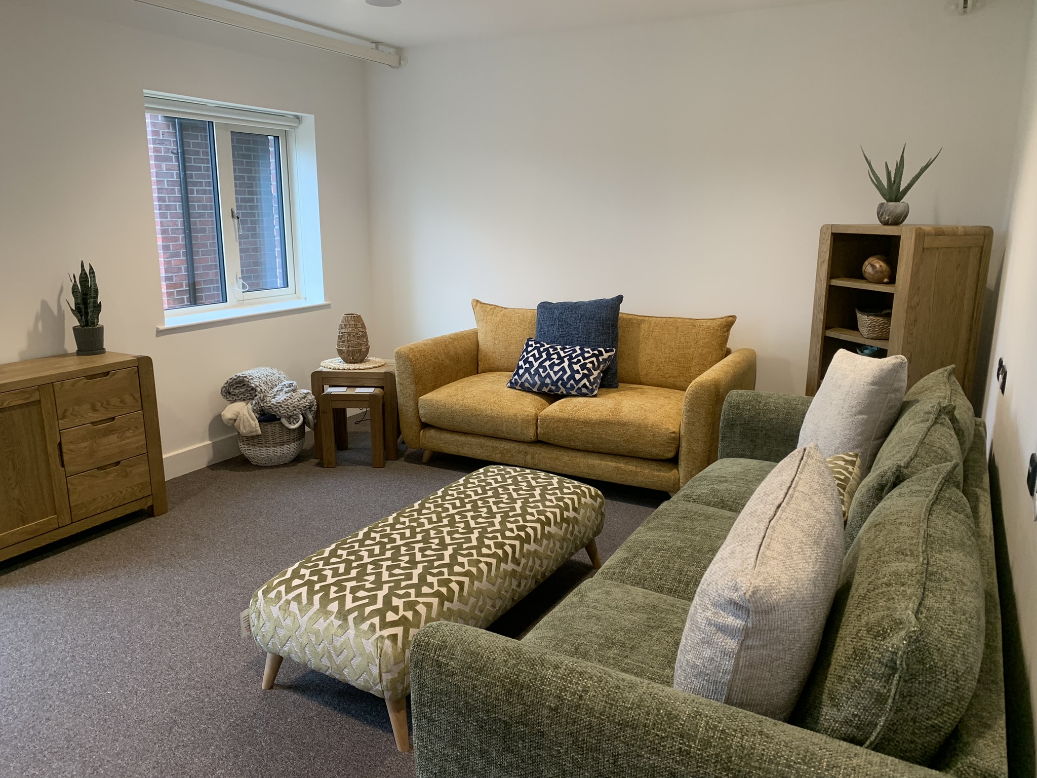 The interior of a children's home in Hull. There's a yellow sofa, some wooden drawers, a green sofa with cushions, a bookshelf, a window and a patterned foot stool.