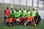A football team crouch together on an indoor pitch in green shirts and red subs vests