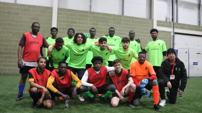 A football team crouch together on an indoor pitch in green shirts and red subs vests