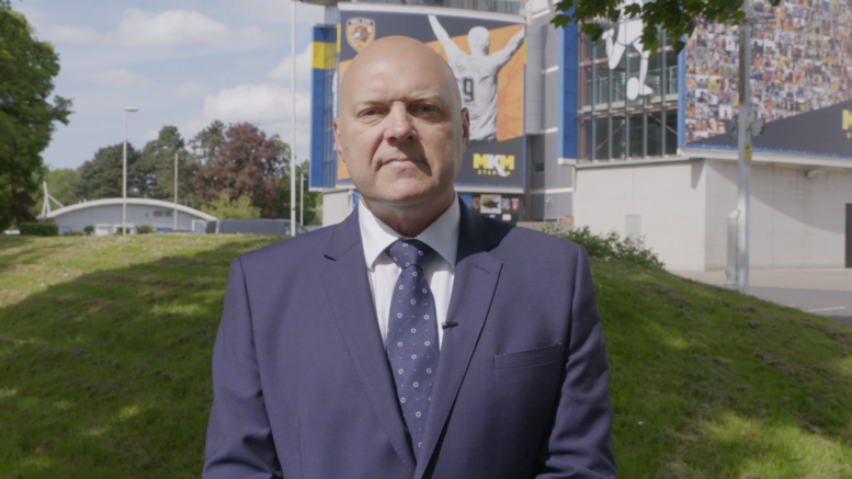 Chris Blacksell stands in a blue suit and tie in front of the MKM Stadium