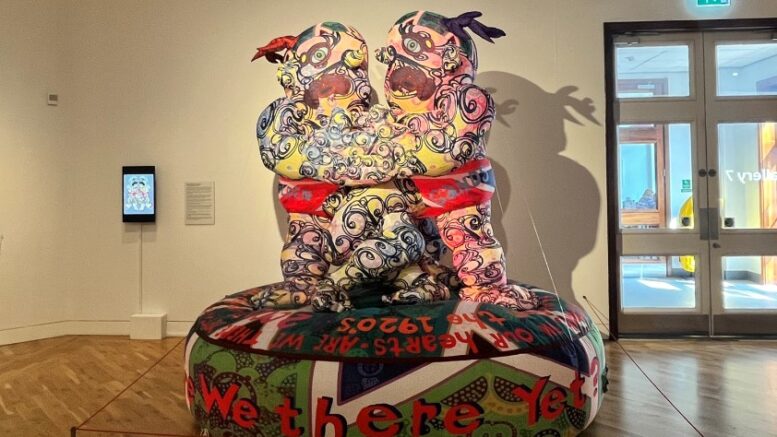 An image of a large, colourful inflatable sculpture of 2 people with tattoo-like markings and intertwined arms, on a platform with 'are we there yet?' written on it.