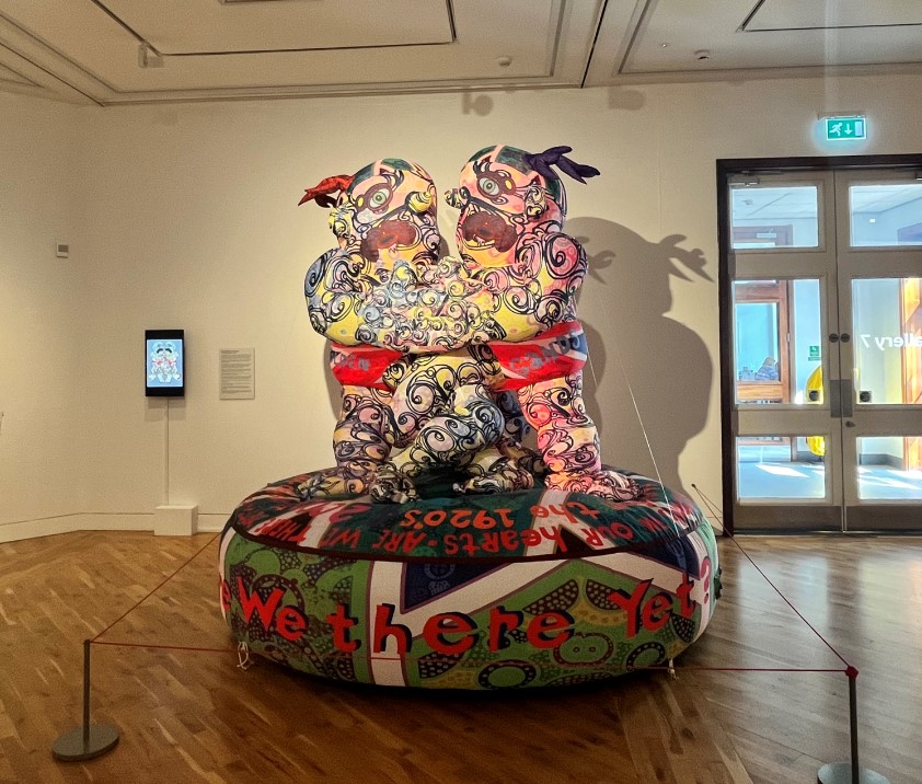 An image of a large, colourful inflatable sculpture of 2 people with tattoo-like markings and intertwined arms, on a platform with 'are we there yet?' written on it.