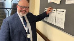 Matt Jukes stands in a blue suit pinning the A4 notices of election to a blue pin board