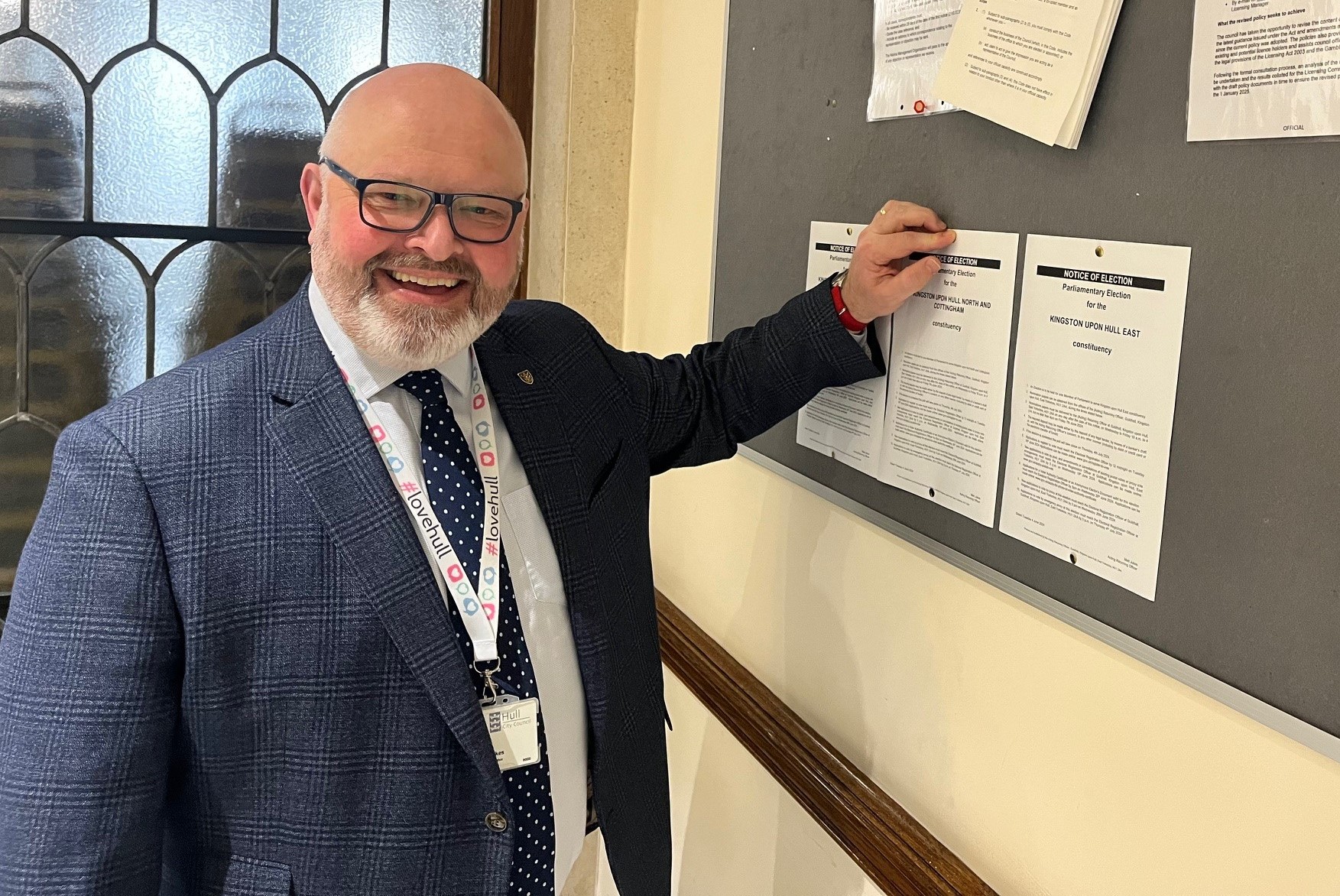 Matt Jukes stands in a blue suit pinning the A4 notices of election to a blue pin board