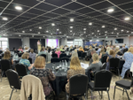 It is a photo of the conference, with people sat at round tables in a large room setting.