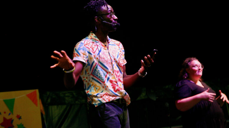 A man with a multicoloured shirt stands on a stage speaking with a female sign language interpreter in the background