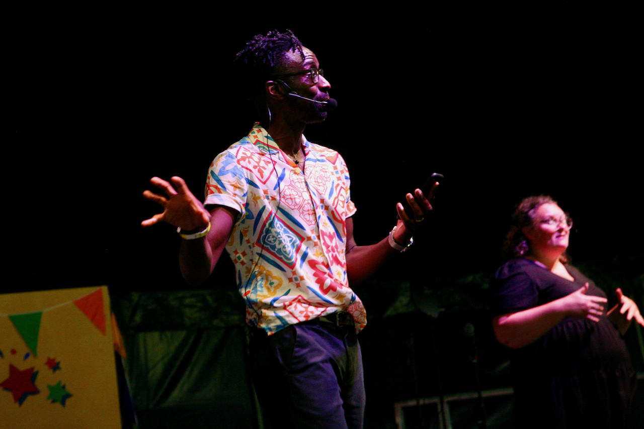 A man with a multicoloured shirt stands on a stage speaking with a female sign language interpreter in the background