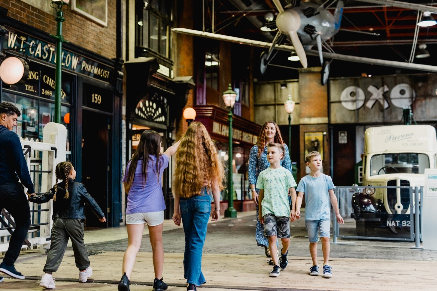 Children and adults walking through the streetlife museum with a vintage car in the background and plane suspended from the ceiling