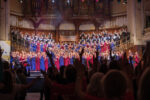 A choir dresses in red and blue robes stand on a stage with silhouettes of cheering crowd in the foreground