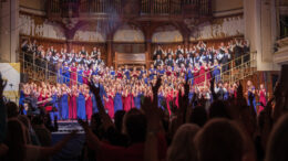 A choir dresses in red and blue robes stand on a stage with silhouettes of cheering crowd in the foreground
