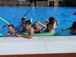 A group of children with pool noodles playing in an outdoor pool in the sun