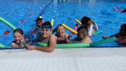 A group of children with pool noodles playing in an outdoor pool in the sun