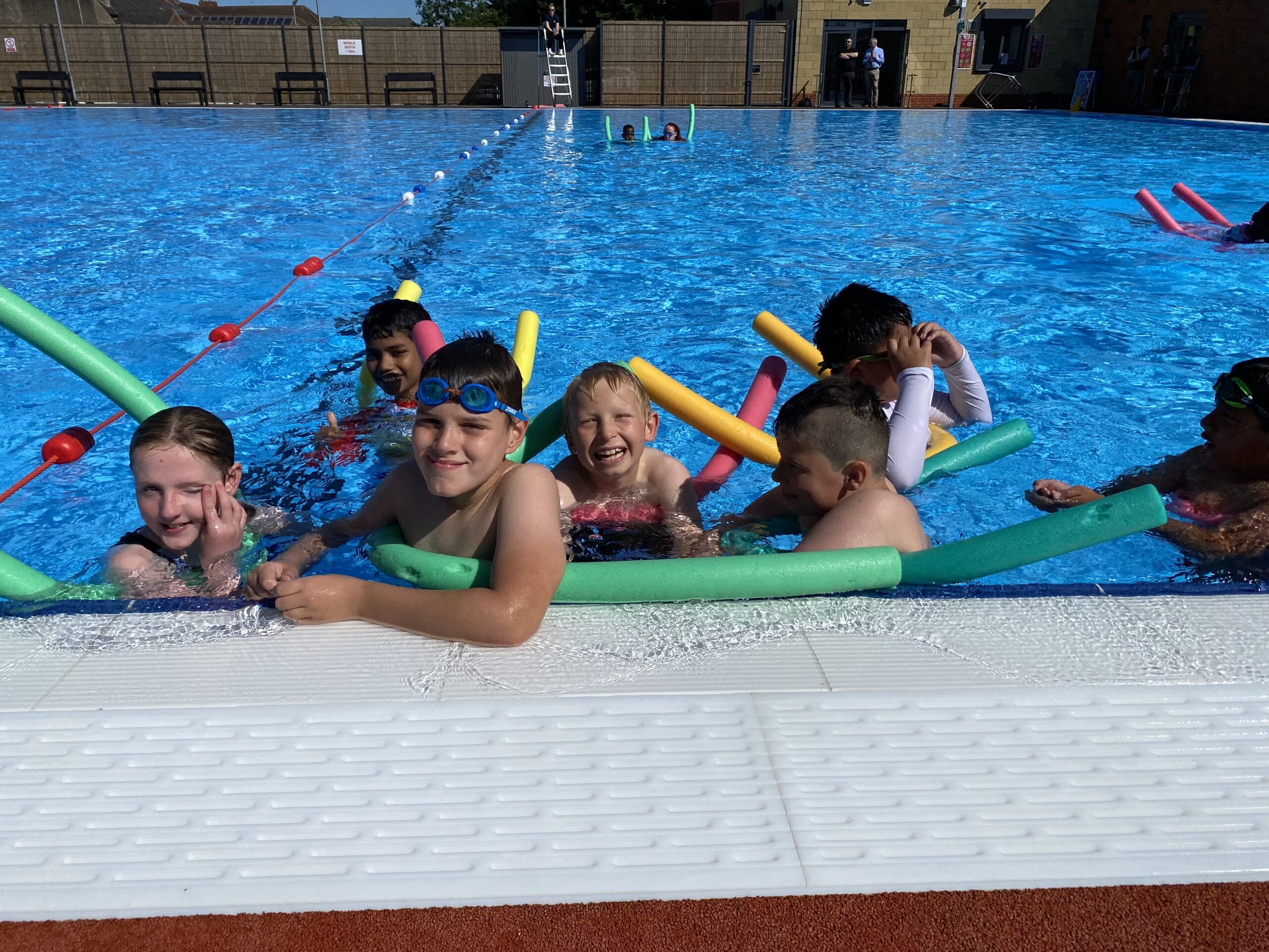 A group of children with pool noodles playing in an outdoor pool in the sun