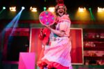 A man dressed as a panto dame on stage holding up a fake pizza to the crowd