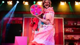 A man dressed as a panto dame on stage holding up a fake pizza to the crowd