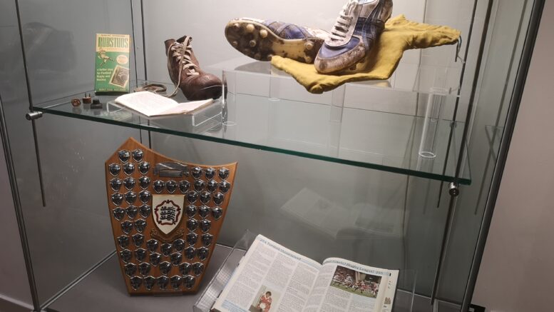 A glass museum case with football boots, a plaque and football programme on display