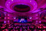 A picture of a large auditorium with circle tables filled with people. Everything lit in pink and purple.