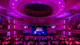 A picture of a large auditorium with circle tables filled with people. Everything lit in pink and purple.