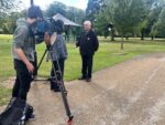 Picture of a man in a black jacket beinf interviewed in a park by a camera crew