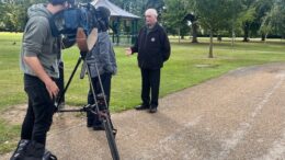 Picture of a man in a black jacket beinf interviewed in a park by a camera crew