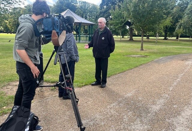 Picture of a man in a black jacket beinf interviewed in a park by a camera crew