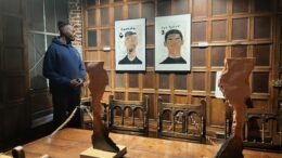 A man in a blue hoody looks at 2 painted portrait pictures hanging on a wooden panelled wall with a wooden sculpture on a table in the foreground