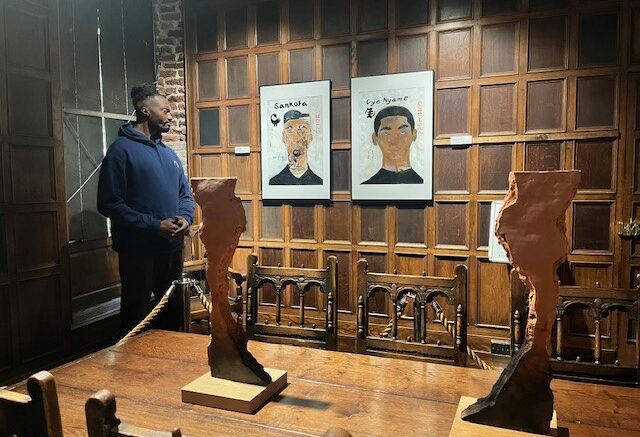 A man in a blue hoody looks at 2 painted portrait pictures hanging on a wooden panelled wall with a wooden sculpture on a table in the foreground