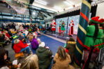 An audience of children and adults watch a stage with Santa, elves and other characters performing.