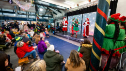 An audience of children and adults watch a stage with Santa, elves and other characters performing.