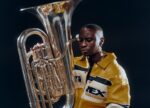 A person with a yellow jacket sits with a silver tuba on their knee