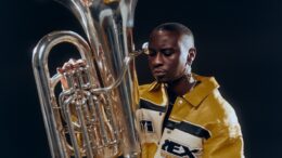 A person with a yellow jacket sits with a silver tuba on their knee