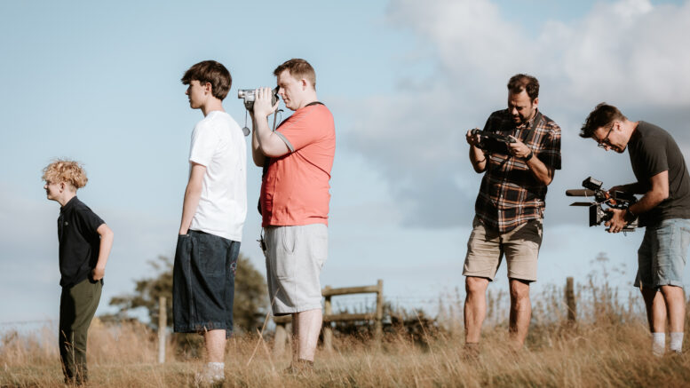 A group of 3 men and 2 young boys with camera equipment. In long grass