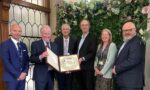 A group of five men and one woman stand in a group. A man in the centre holds open a case with a large silver award inside. They are in a brightly lit room, with panelling and a floral display behind them