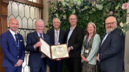 A group of five men and one woman stand in a group. A man in the centre holds open a case with a large silver award inside. They are in a brightly lit room, with panelling and a floral display behind them