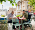 2 people sit at an outdoor dining table in a city centre. one is in a wheelchair