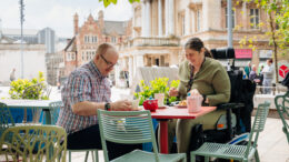 2 people sit at an outdoor dining table in a city centre. one is in a wheelchair