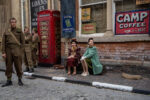 A vintage style movie set with two women in 30s clothing sat on a path next to a red phone box and three soldiers in uniform.