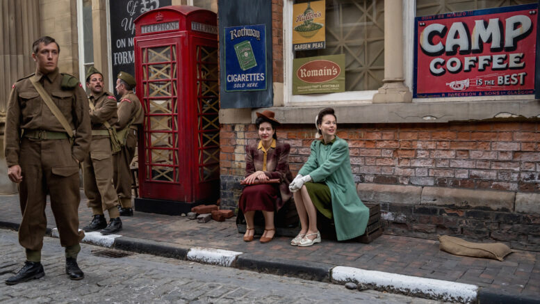 A vintage style movie set with two women in 30s clothing sat on a path next to a red phone box and three soldiers in uniform.