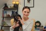 A young brunette woman sits on a chair in a living room set up holding a black and brown Dachshund