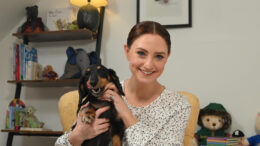 A young brunette woman sits on a chair in a living room set up holding a black and brown Dachshund