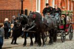 Tow black horses on a cobbled street pulling a black and red carriage with two drivers in Victorian Clothing