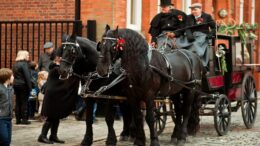 Tow black horses on a cobbled street pulling a black and red carriage with two drivers in Victorian Clothing