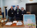 three people stand behind a stall with leaflets and pens. There is a bright A Board with a poster advising people about 'Neighbourhood Nuisance'