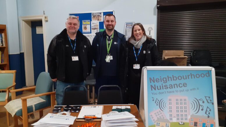 three people stand behind a stall with leaflets and pens. There is a bright A Board with a poster advising people about 'Neighbourhood Nuisance'