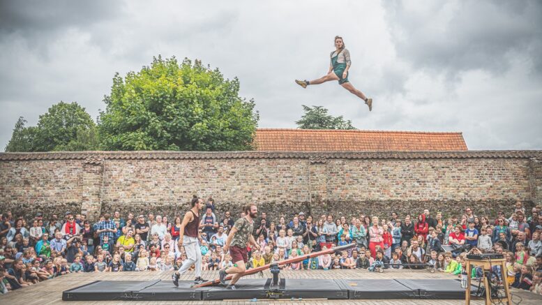 A crowd watch as a person is launched into the air from a seesaw on the ground