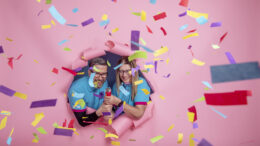 two people in blue volunteer tshirts bursting through a pink wall with a large confetti cannon