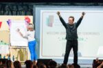 A man stood on a stage with his arms raised in the air with a woman stood behind him drawing on a flip chart