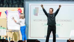 A man stood on a stage with his arms raised in the air with a woman stood behind him drawing on a flip chart