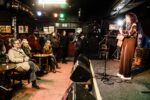 A female singer performs on a stage with a guitar in a dark venue with an audience watching