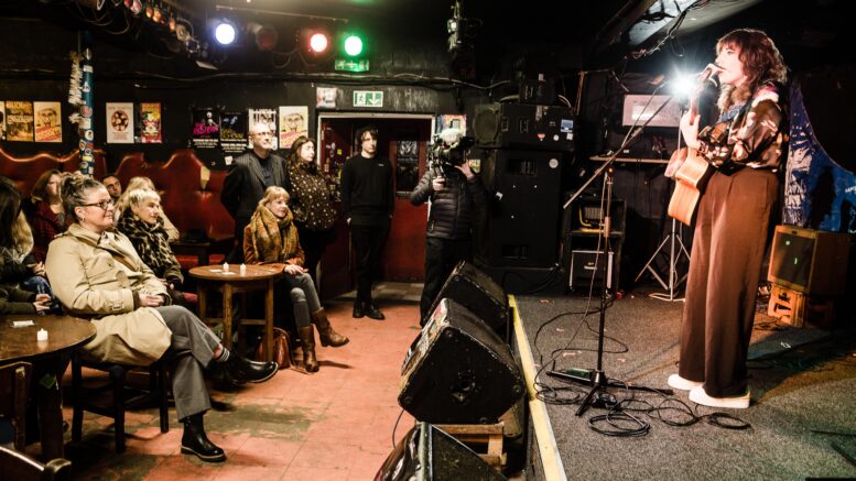 A female singer performs on a stage with a guitar in a dark venue with an audience watching